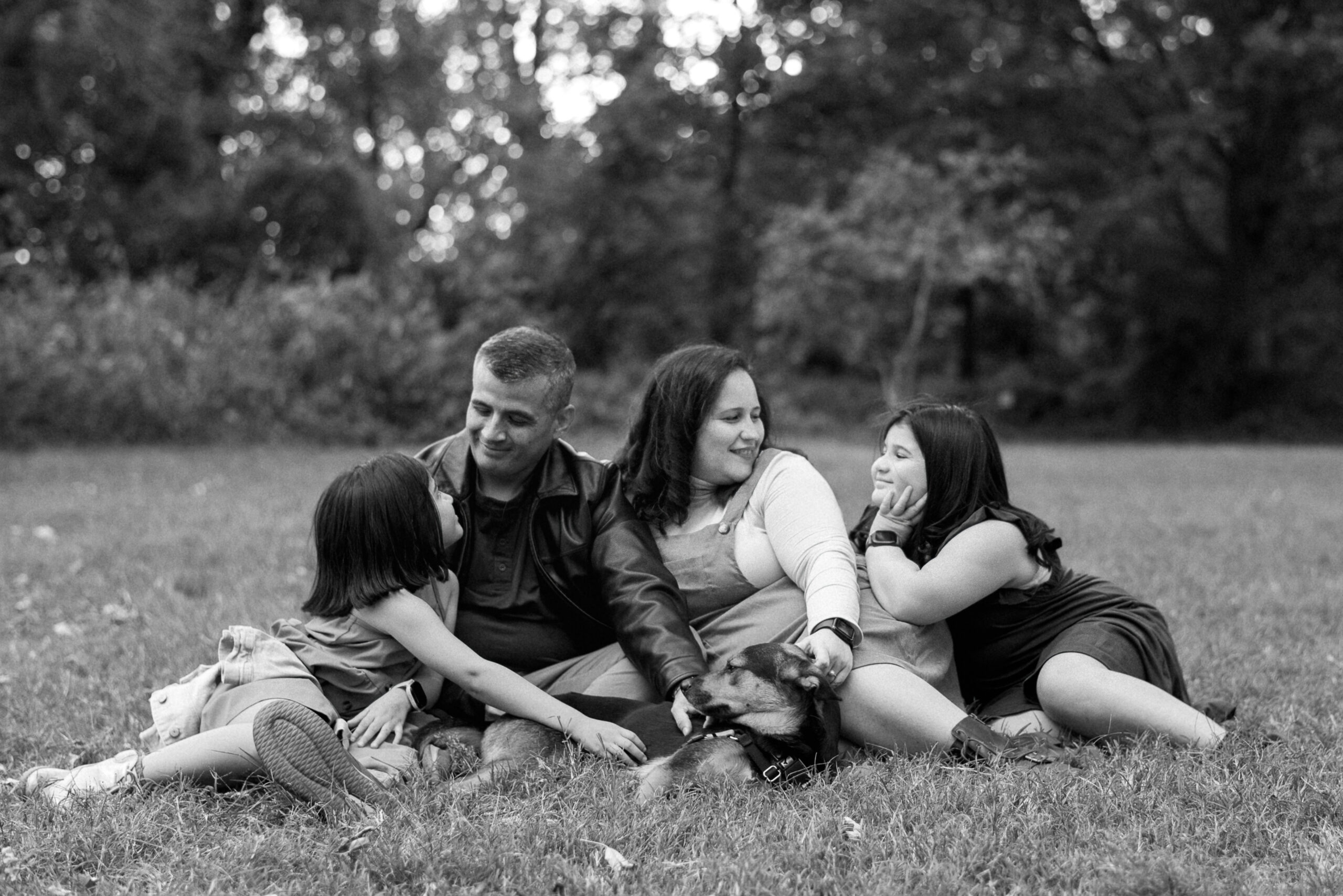 black and white image of family of 4 with two young girls sitting together in grass with puppy