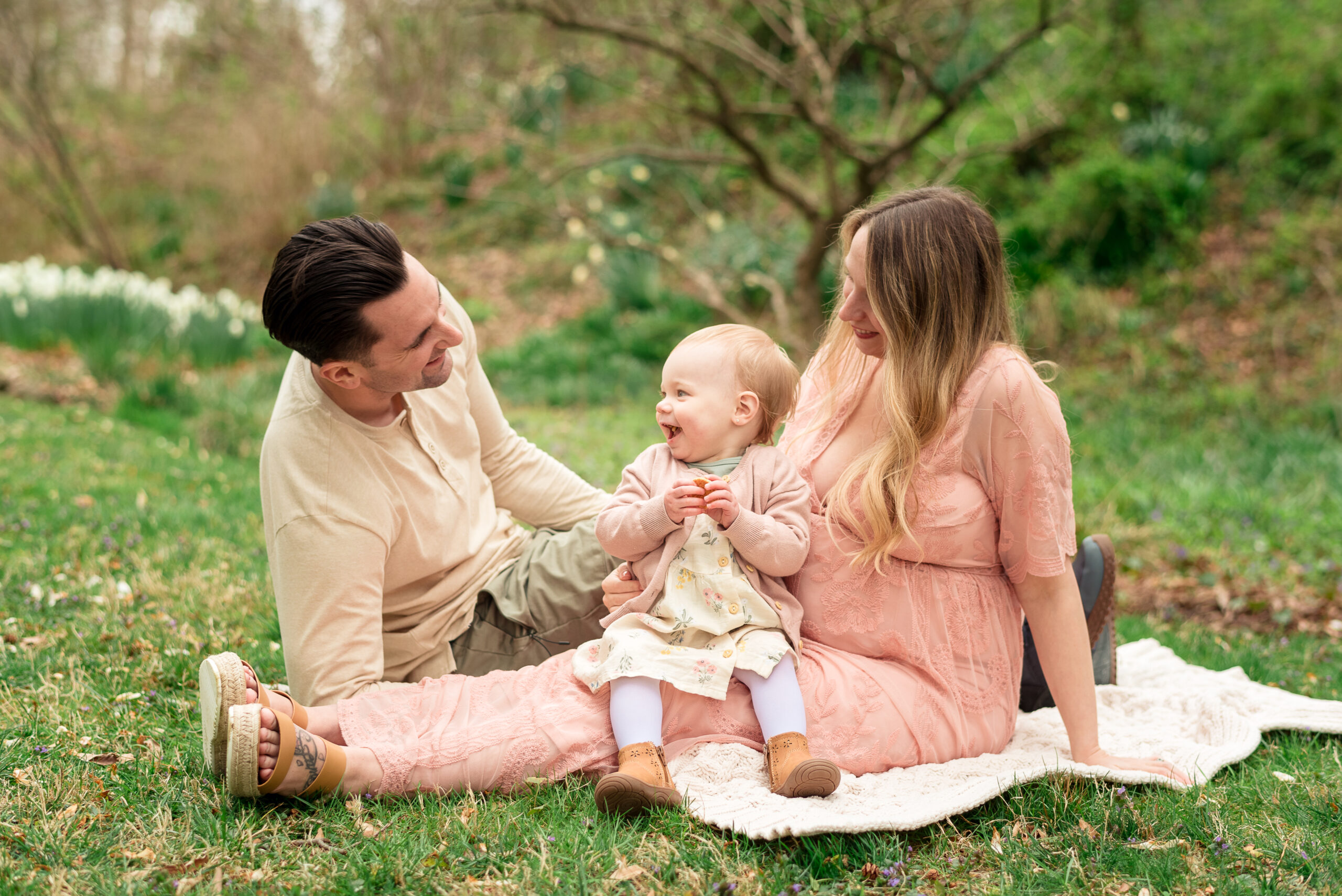 pregnant mom with toddler on lap and husband sitting across on blanket in field