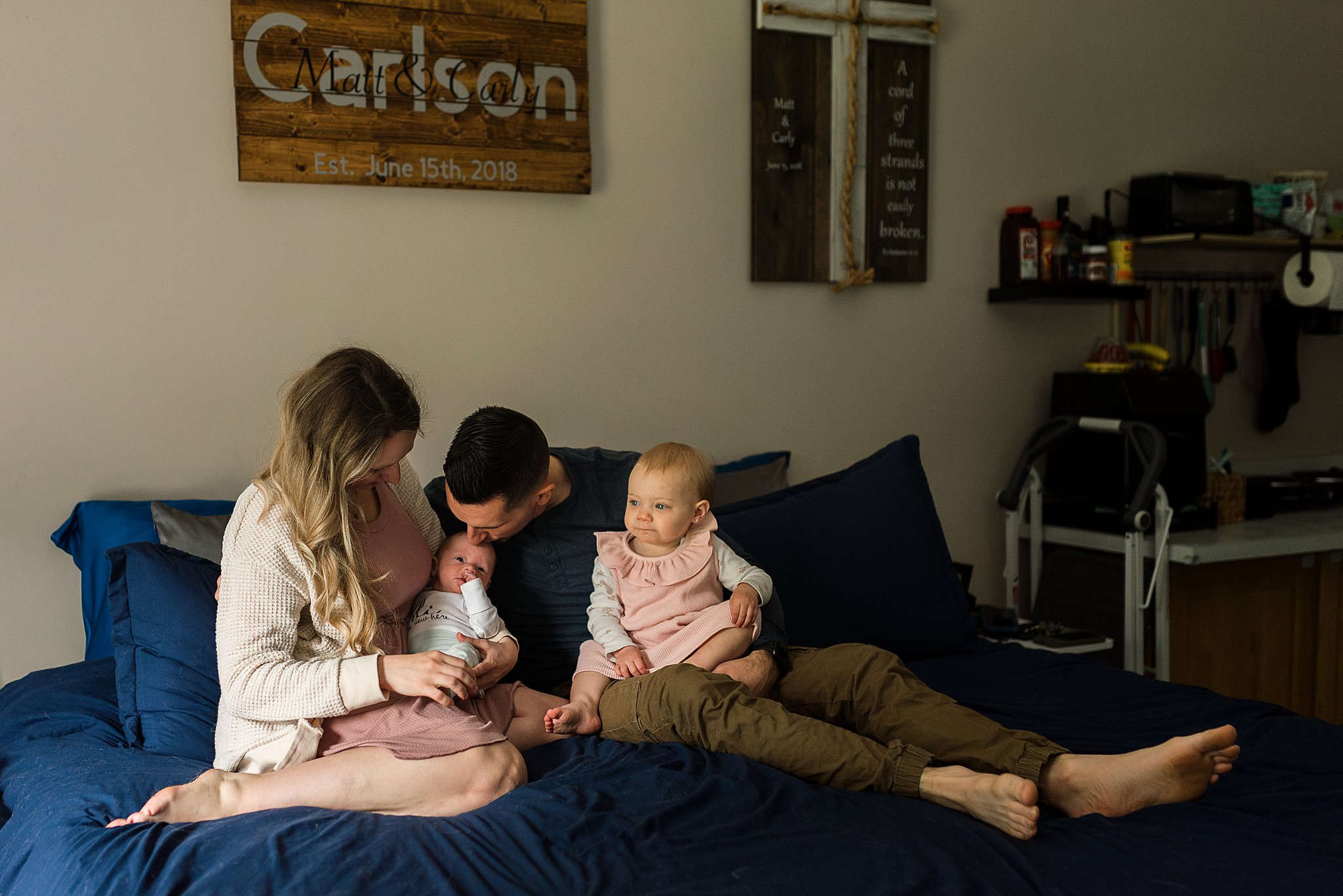 family of 4 sitting on bed, dad kissing newborn